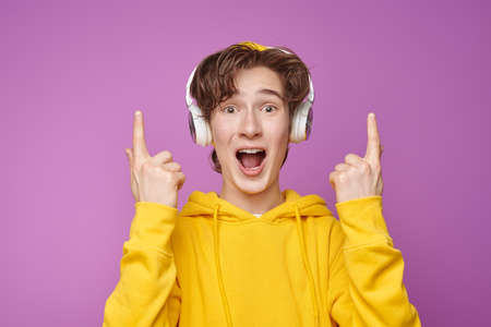 A cool modern teenager in stylish bright youth clothes and headphones smiles and joyfully points his index finger up. Purple background. Youth lifestyle and emotions.の写真素材
