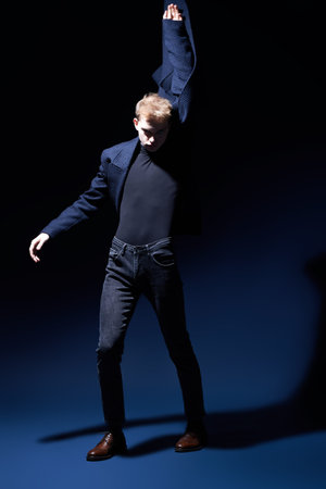 Studio shot of a young blond man in blue coat, turtleneck and jeans looking aggressively into the...