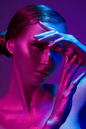 Art portrait of a beautiful, feminine fashion model with shiny silver skin in colorful neon blue and red light on a purple background. Beauty and cosmetics concept. Body painting.の写真素材