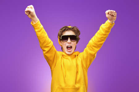 Cool teenage boy in bright yellow hoodie and black sunglasses rejoices emotionally clenching his fists. Bright purple background. Youth lifestyle, success. Adolescents and emotions.の写真素材