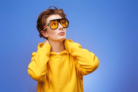 Adolescents, emotions. Portrait of a calm modern teenage boy in bright yellow hoodie holding his hands behind his head and looking pensively up. Blue background at studio.の写真素材