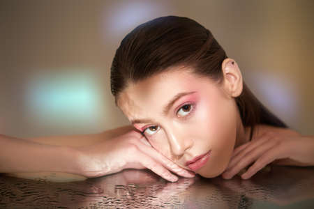 Beautiful model woman with light makeup looks calmly at the camera bending down her face over clean water. Studio shot. Women's beauty and health. Cosmetics and skin care.の写真素材