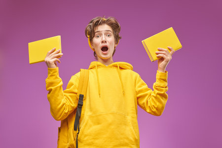 A modern teenager in bright youth clothes with a backpack and books looking with surprise and sadness into the camera. Purple background. Educational concept.の写真素材