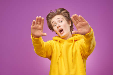 Youth lifestyle and emotions.  A funny modern teen boy wearing stylish bright yellow hoodie looks at the camera through his hands. Purple background.の写真素材