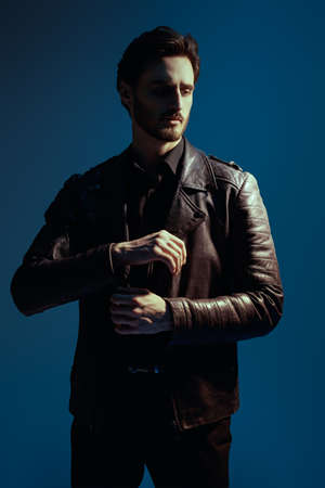 Men's beauty, fashion. A handsome brutal brunet man in a leather jacket poses on a dark blue background.の写真素材