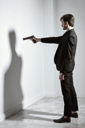 Portrait of a brunet man in a black suit standing in front of the the wall  aiming with a gun in his...
