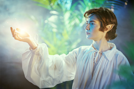 Fairytale young elf looks at the magic light in his hand while standing in the forest. Fairy tale,...