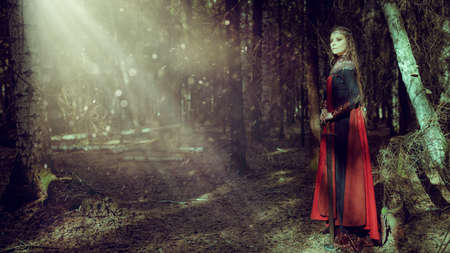 A beautiful medieval queen stands with a sword in a dark dense forest with the light breaking through the branches of the trees. Ancient legends. Fantasy world. Copy space.の写真素材