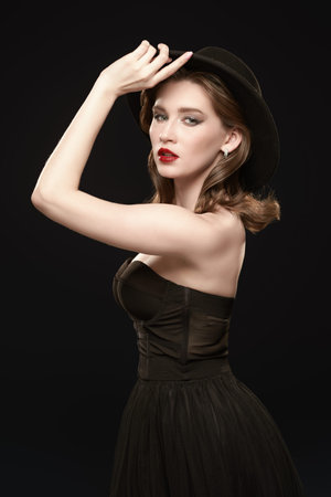 Adorable brunette girl with bright evening makeup poses in elegant black dress and hat on a black...