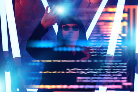 Teen boy hacker. A modern teen boy in a black gown, mask and black sunglasses hiding his face works with hologram in a dark space among neon lights. Futuristic technologies.の写真素材