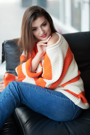 A pretty smiling girl having a rest in her cozy living room, sitting on a leather sofa. Home...