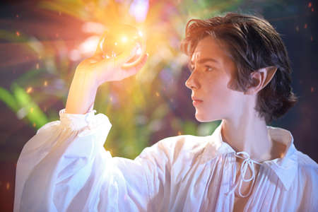 Courageous fairytale elf boy holds a magic sphere of light in his hand while standing in the forest. Fairy tale, magic. Fantasy.の写真素材