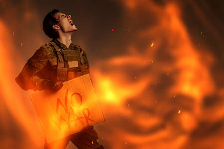 No War concept. Angry soldier in Protective Combat Uniform holds a sign "No War" screaming furiously in a dark background engulfed in flames. Studio shot.の写真素材