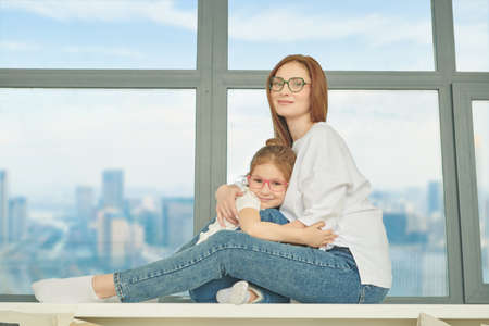 Mother and her little daughter sit on a windowsill in modern apartment hugging and joyfully smiling. Happy family.の写真素材