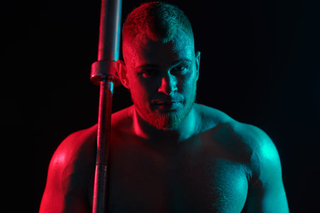 A handsome male weightlifter does exercises with a barbell on a dark studio background with crimson...