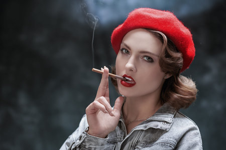 Attractive confident girl in denim overalls and a red beret smokes a cigarette. Studio portrait on a...