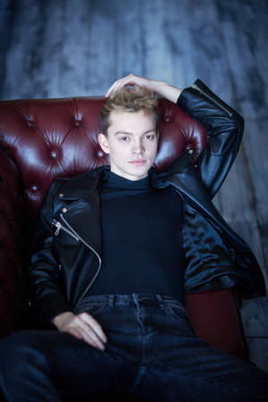 A stylish young man in black leather jacket and black jeans rests on a leather sofa in loft style room. Men's fashion and style.の写真素材