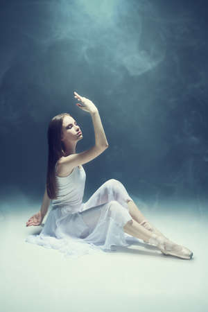 Ballet performance. Beautiful  ballerina in a light, white dress and pointe shoes dances gracefully, surrounded by smoke. Full-length studio portrait. Art of dance, modern ballet.の写真素材