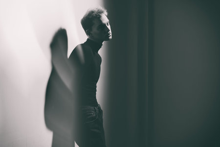 Portrait of a sad young man in a black turtleneck and jeans leaning against a white wall and looking...