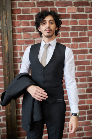 A handsome smiling young man in an elegant black suit and a white shirt stands by a brick wall....