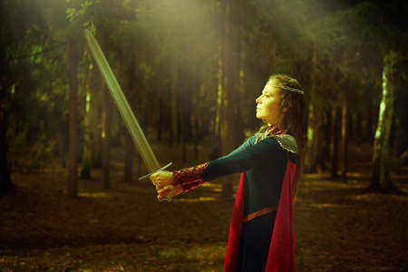 Beautiful medieval woman warrior looks at the sword in her hands standing in a dense forest in sunlight. Historical reconstruction. Legends of Valiant Knights. Fantasy.の写真素材