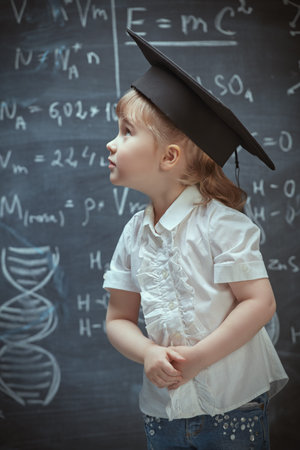 Kids and education. A smart little girl in white shirt and academic cap stands by the blackboard with formulas and diagrams. Educational concept.の写真素材
