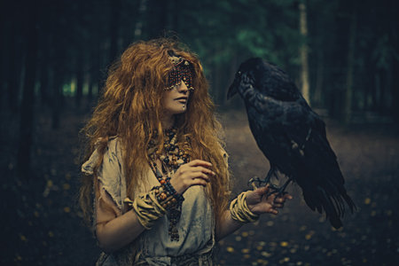 Halloween. A fairytale forest witch with a mask covering her eyes talks to a black raven, standing...