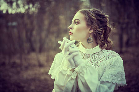 Vintage portrait of a beautiful noble lady in a white dress standing in the old park. Hair and makeup, late 19th century. Historical reconstruction of the Victorian era.の写真素材