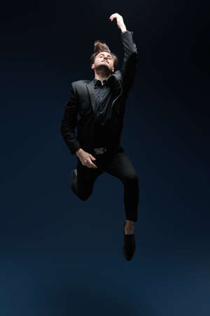 A man in a black classic suit jumps, feeling freedom and lightness. Studio full-length portrait on a dark blue background. Happiness. The concept of success and aspirations, dreams.の写真素材