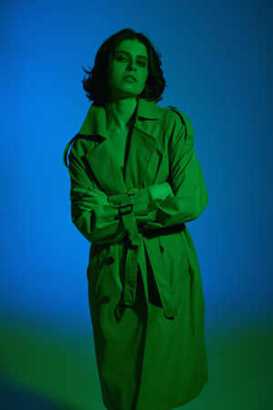Beautiful brunette girl model posing at the studio in a raincoat on a blue and green background in green lighting. Fashion shot.の写真素材