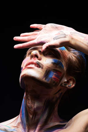 Body painting, art. Portrait of a handsome blond man with colorful painted face posing artistically with his hand on a black background.の写真素材