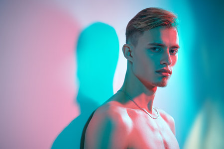 Studio shot of an attractive young man with naked athletic torso posing on a light background in multicoloured lighting. Men's beauty and health. Copy space.の写真素材