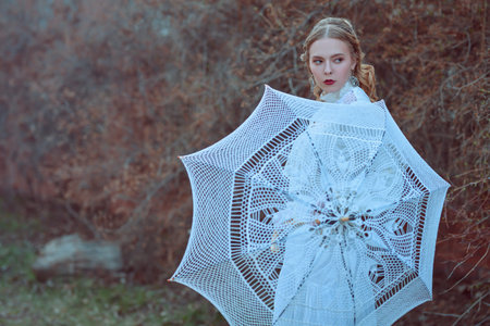 Vintage portrait of a stunning aristocratic girl in a white lace dress walking outdoors holding a...