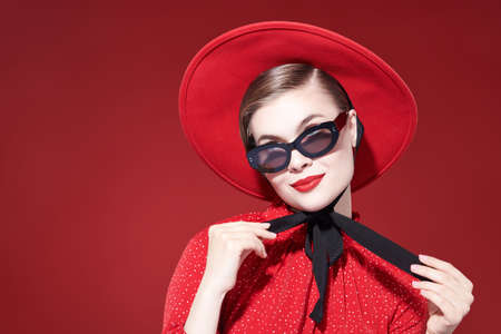 A happy fashionable young woman with bright makeup poses in a red dress, red hat and stylish sunglasses and smiles charmingly.  Red background. Beauty, makeup and cosmetics. Fashion.の写真素材
