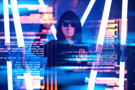 Teen boy hacker. A modern teen boy in a black gown, mask and black sunglasses hiding his face works with hologram in a dark space among neon lights. Futuristic technologies.の写真素材