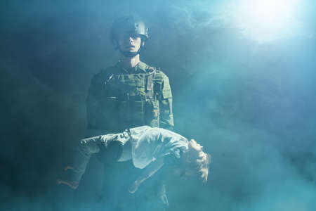 Portrait of a soldier carrying a little injured girl in his arms. Dark background shrouded in smoke. Casualties of war.の写真素材