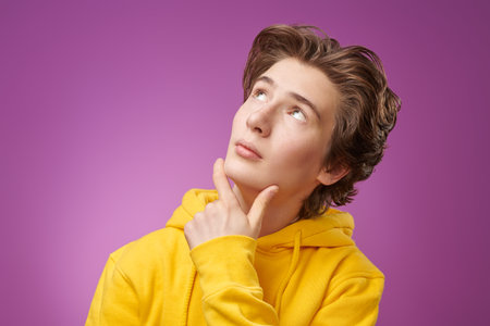 Portrait of a calm modern teenage boy in bright yellow hoodie rubbing his chin and looking pensively...