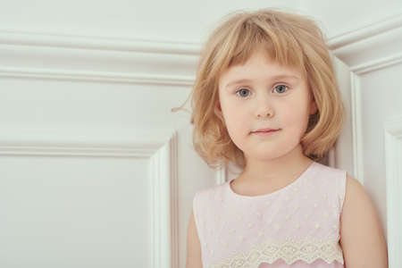Happy little child girl with blonde hair looks calmly with slight smile at the camera. Studio portrait in a white classic apartment. Happy childhood. Copy space.の写真素材