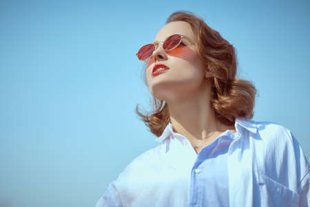 Beautiful brunette woman with elegant makeup, hairstyle and sunglasses wearing a blue shirt stands outdoors on a sunny spring day in the background of blue sky. Makeup, hairstyle and glasses style.の写真素材