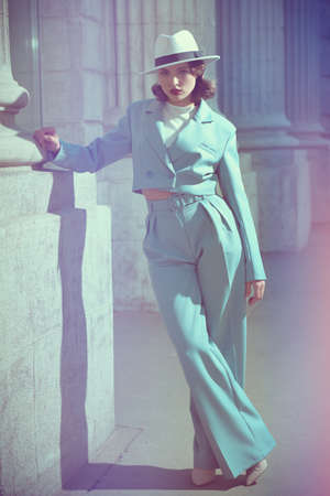 Portrait of a beautiful brunette woman in an elegant sky blue pantsuit and a white hat, standing by a building on a city street. Style of the 40s. Makeup and hairstyle. Hollywood style.の写真素材