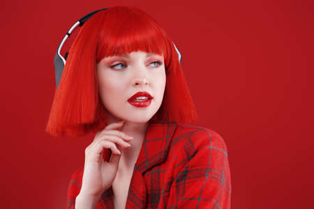 DJ, party girl. A stylish girl with bright red lips and red hair wearing a red checkered jacket and headphones. Red studio background. Beauty, Fashion.の写真素材