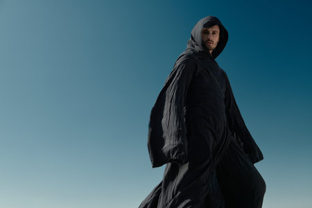 A brave man in a black robe with a hood on his head stands on top of a mountain against a blue sky, blown by the winds. Wanderer, pilgrim. Adventure and Fantasy story. Copy space.の写真素材