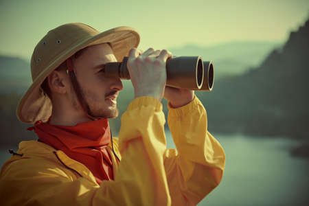 Tourism and travel. A man traveler-explorer looks into the distance through binoculars against the backdrop of a mountain landscape. Discoverer. Retro style.の写真素材