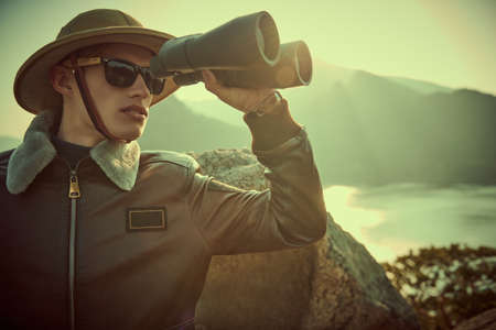 A courageous male discoverer in a leather jacket and hat looks through binoculars in search of new adventures. Mountain landscape and sunset in the background. Retro style.の写真素材