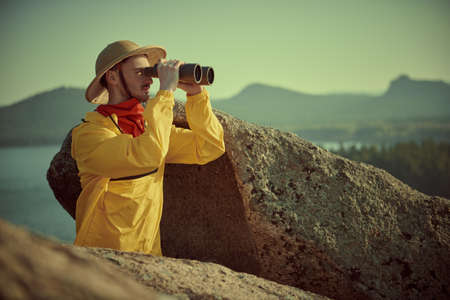 Male adventurer, tourist looks into the distance through binoculars, standing in the mountains. Tourism and travel. Discoverer. Retro style.の写真素材