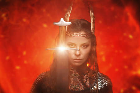 Scandinavian mythology. A beautiful woman valkyrie in a winged helmet holds a sword in front of her in the background of burning fire.の写真素材