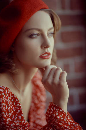 A beautiful young woman with elegant makeup and hairstyle poses in a red dress and red beret. Brick wall background. Beauty, fashion. Elegant retro girl.の写真素材