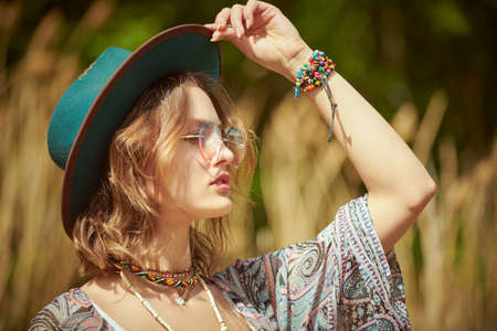 A beautiful romantic girl in boho style clothes and accessories stands in a field near the reeds and looks into the distance. Boho and hippie style. Summer, vacation.の写真素材