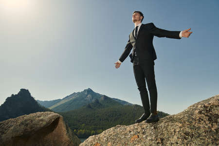 The concept of success. A purposeful businessman in a suit stands on top of a mountain with his arms spread out to the sides and looks into the distance.の写真素材
