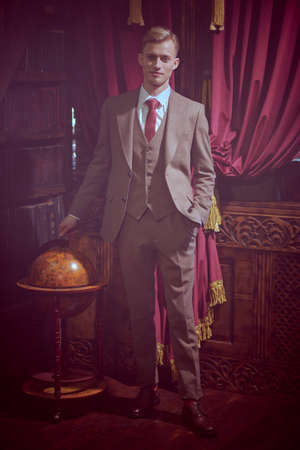 Luxury lifestyle. A well-groomed respectable young man in an expensive coat and three-piece suit standing in luxury apartments with vintage interior. Men's beauty, fashion.の写真素材